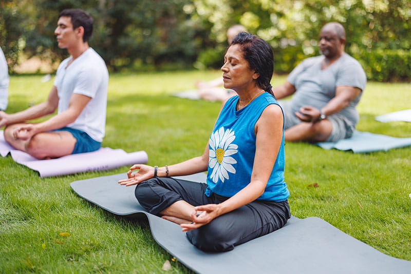A group of people practicing yoga outside, sitting with legs crossed on yoga mats in the grass